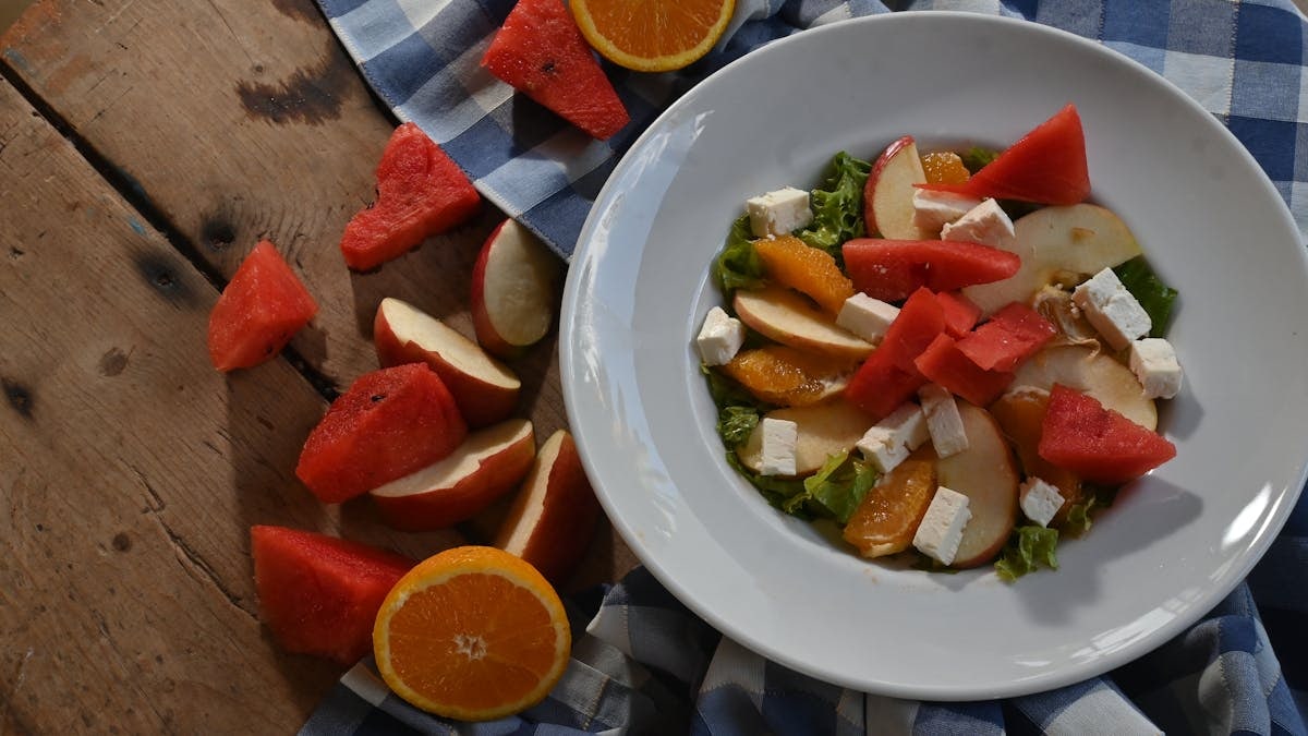 Colorful summer salad featuring fresh watermelon, orange slices, apple slices, and feta cheese on a rustic table.